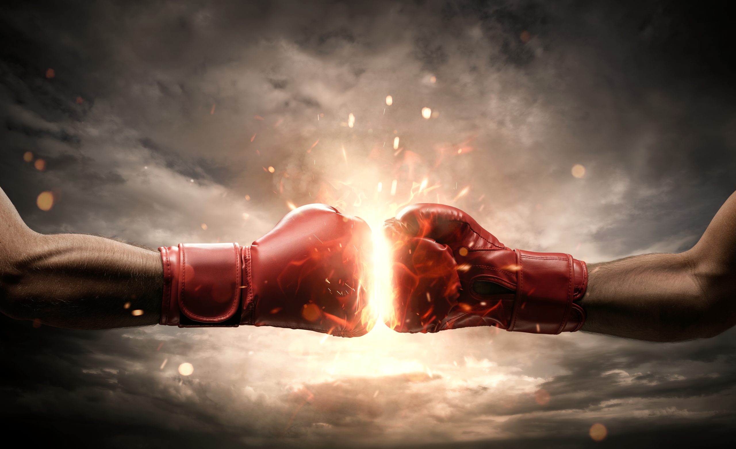Two red boxing gloves striking each other head on with sparks and light at the point of impact, against a dark, dramatic sky
