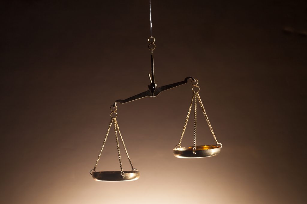 A set of gold scales of justice hanging against a dark background with one pan resting low and the other raised