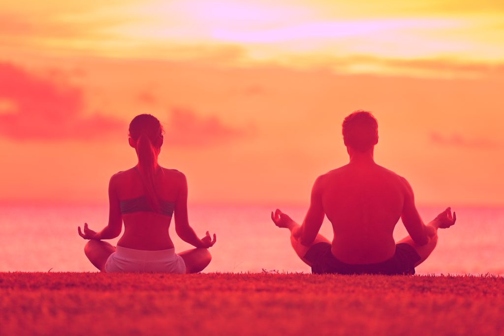 Two people seated in a meditation posture on the grass above the ocean at sunrise