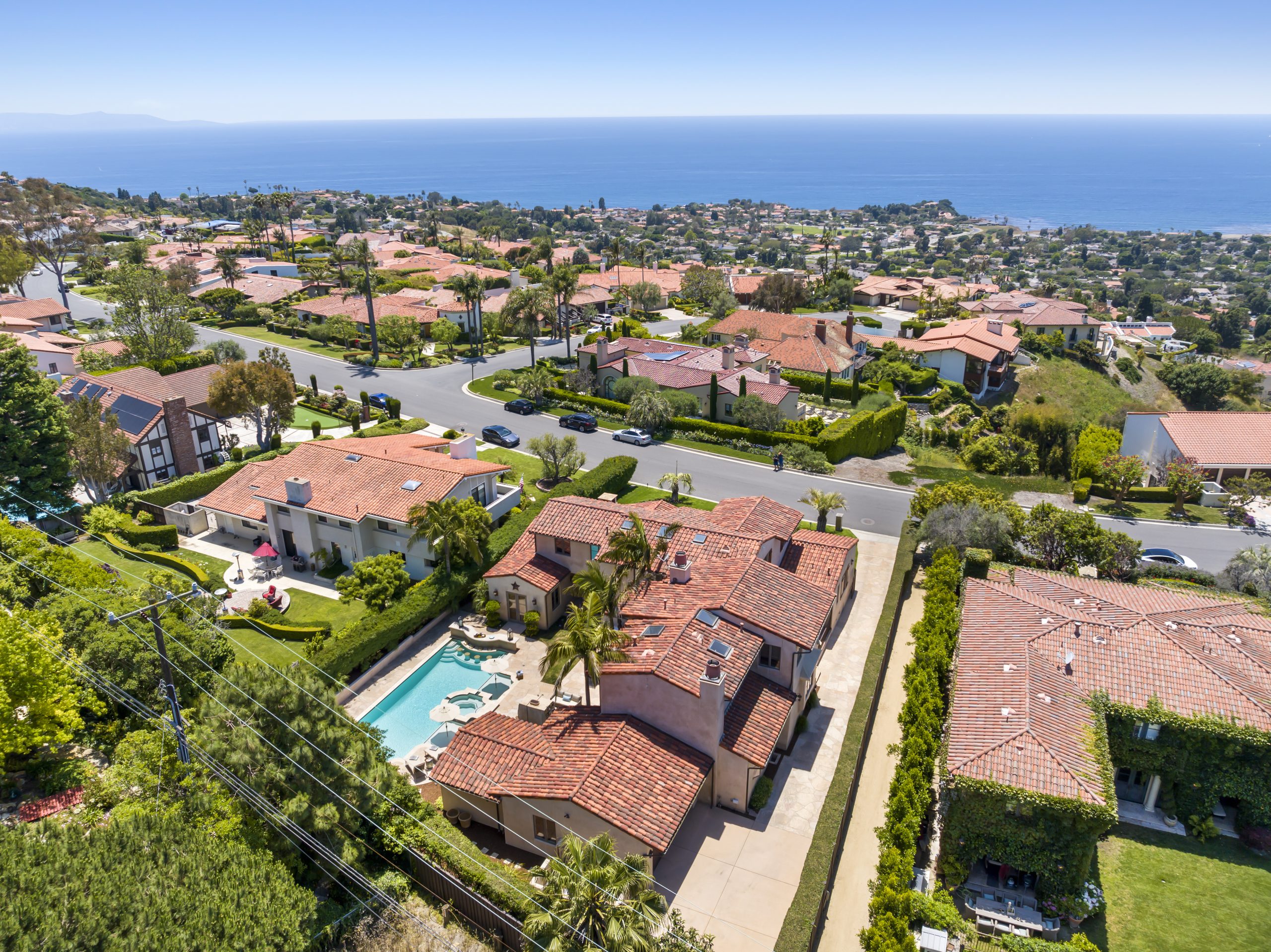 Aerial over a hillside neighborhood of tile-roofed homes with pools and the ocean beyond