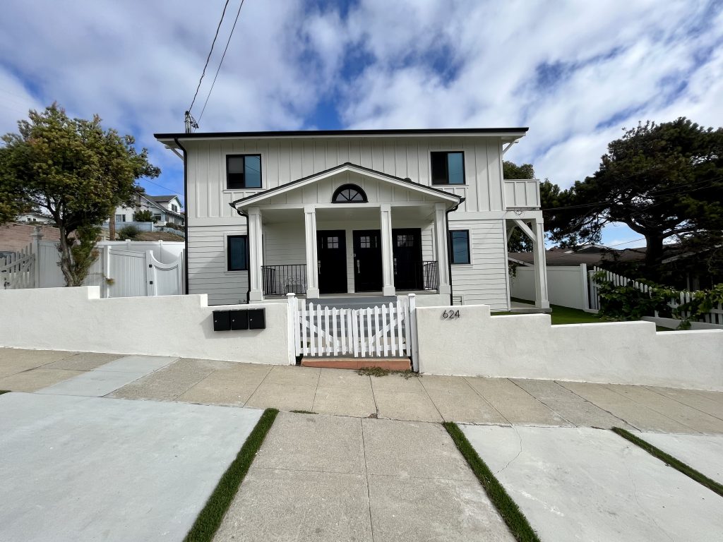 White two-story board and batten building at 624 W. 38th Street in San Pedro, with black-framed windows, a covered front porch with three black doors, and a white picket gate set in a low stucco wall.
