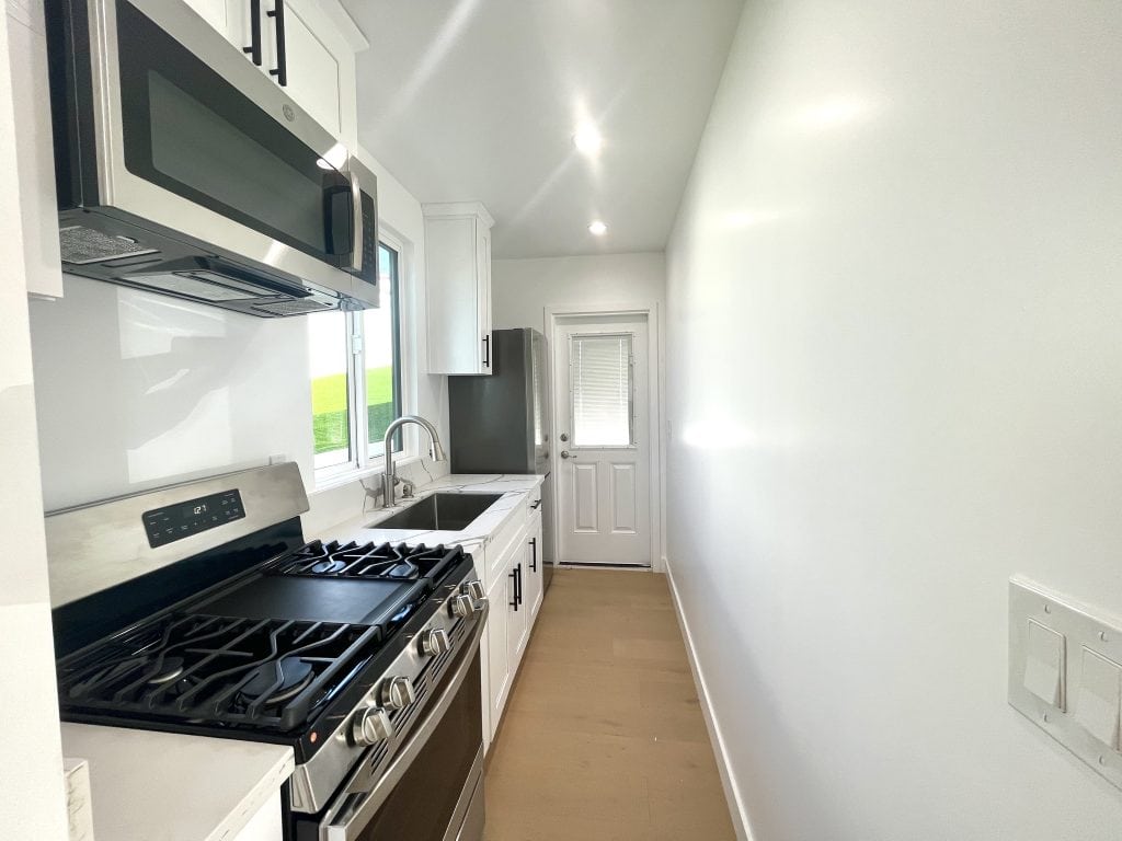 Galley kitchen in Unit A at 624 W. 38th Street, San Pedro, with a stainless gas range and microwave, white shaker cabinets with black pulls, a sink under a window, a stainless refrigerator and a door at the far end.