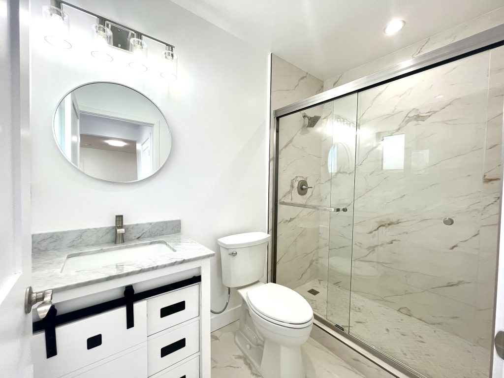 Bathroom in Unit A at 624 W. 38th Street, San Pedro, with a white vanity with black barn-door hardware, a round mirror, and a walk-in shower in marble-look porcelain tile behind a sliding glass door.
