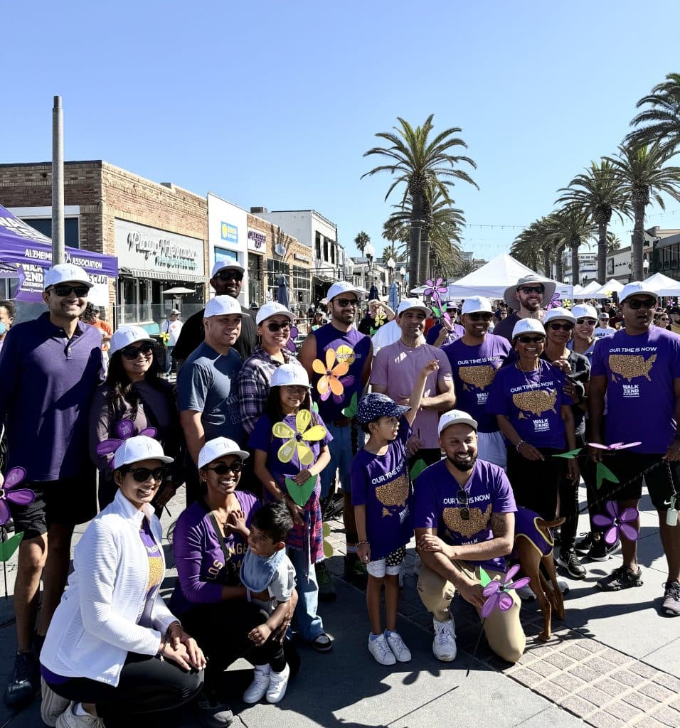 About twenty adults and children in purple Walk to End Alzheimer's shirts and white caps, holding purple, yellow and blue Promise Garden flowers on Pier Avenue in Hermosa Beach, with event tents and palm trees behind them.
