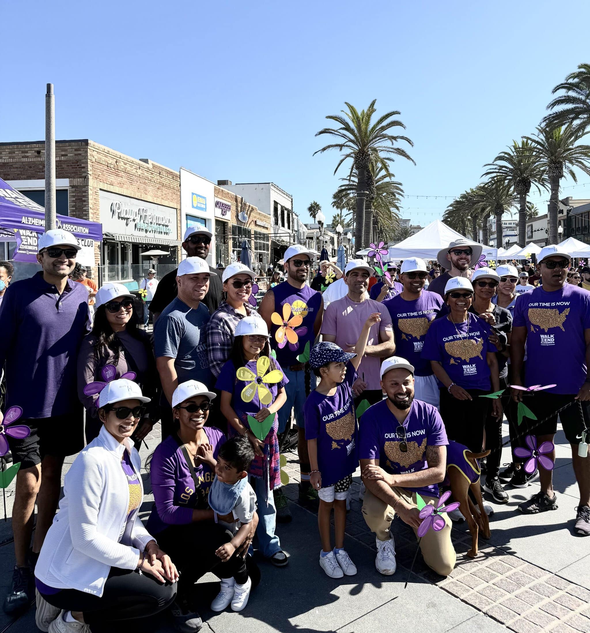 About twenty adults and children in purple Walk to End Alzheimer's shirts and white caps, holding purple, yellow and blue Promise Garden flowers on Pier Avenue in Hermosa Beach, with event tents and palm trees behind them.