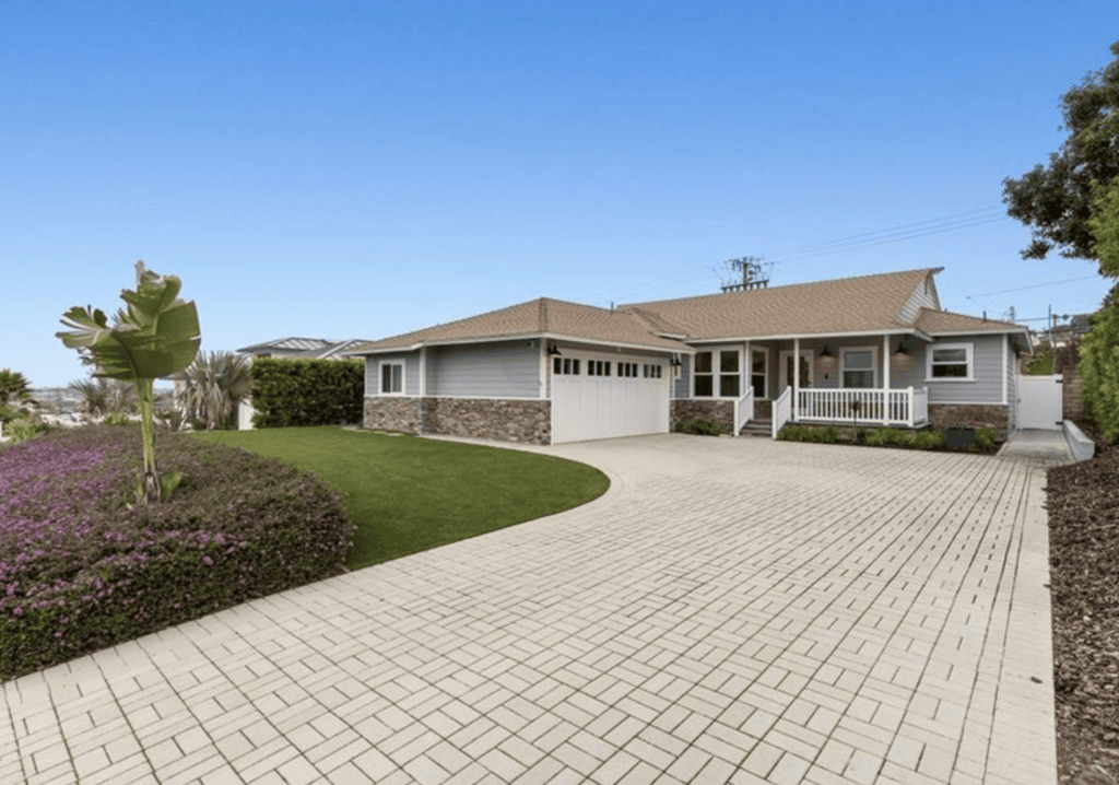 Single-story home with a wide paved driveway, a low hedge and the ocean on the horizon