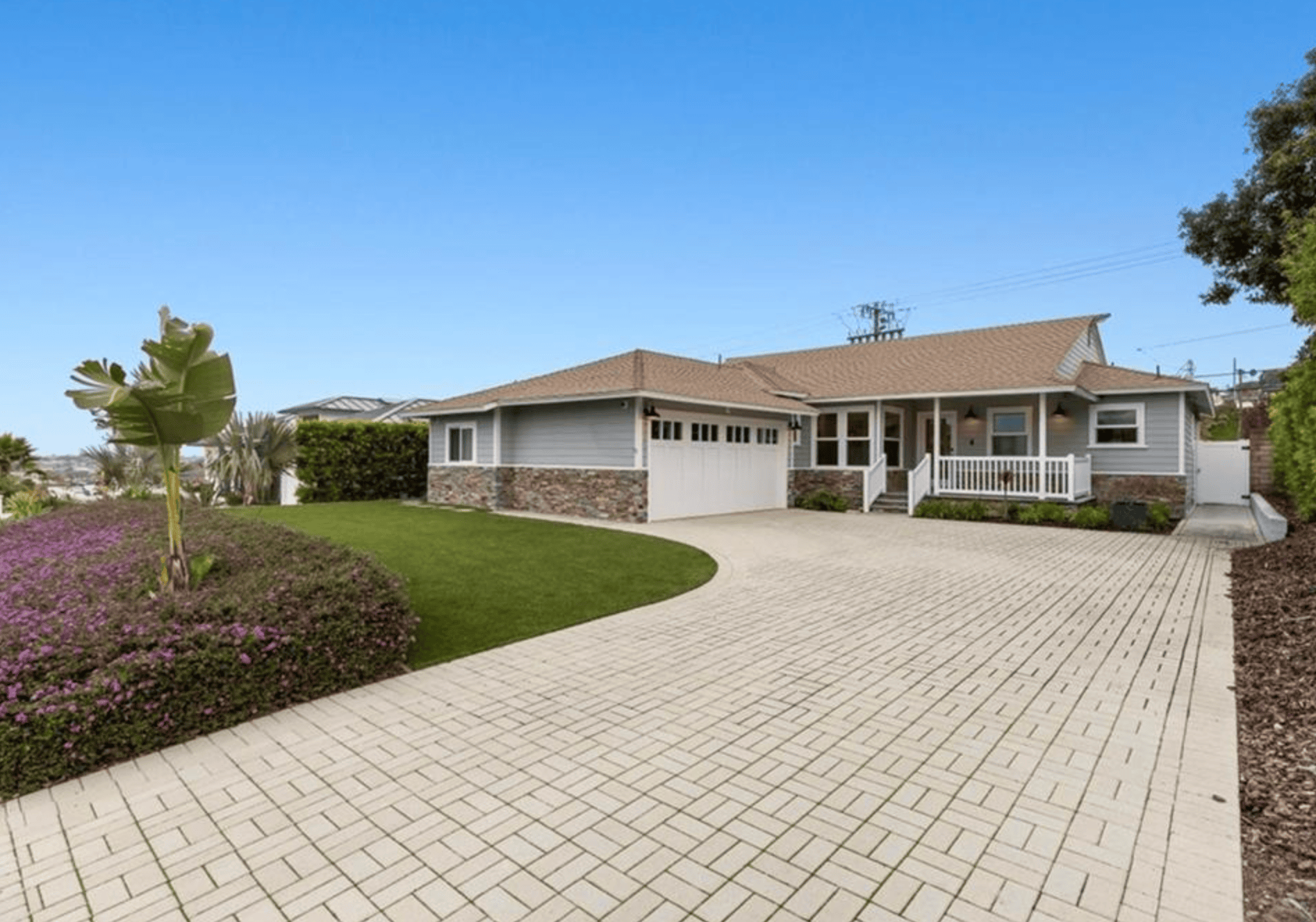 Single-story home with a wide paved driveway, a low hedge and the ocean on the horizon