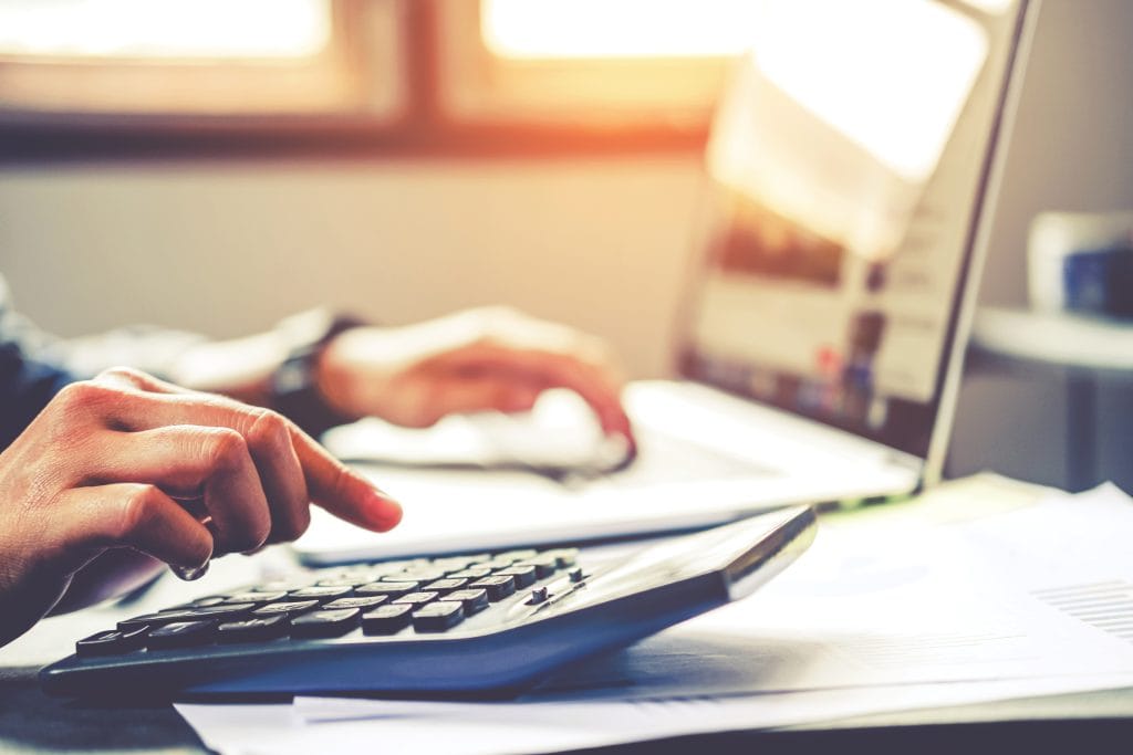 Two hands at a desk, one pressing keys on a desktop calculator and the other resting on a laptop keyboard, with printed documents on the desk beside them and a bright window behind.