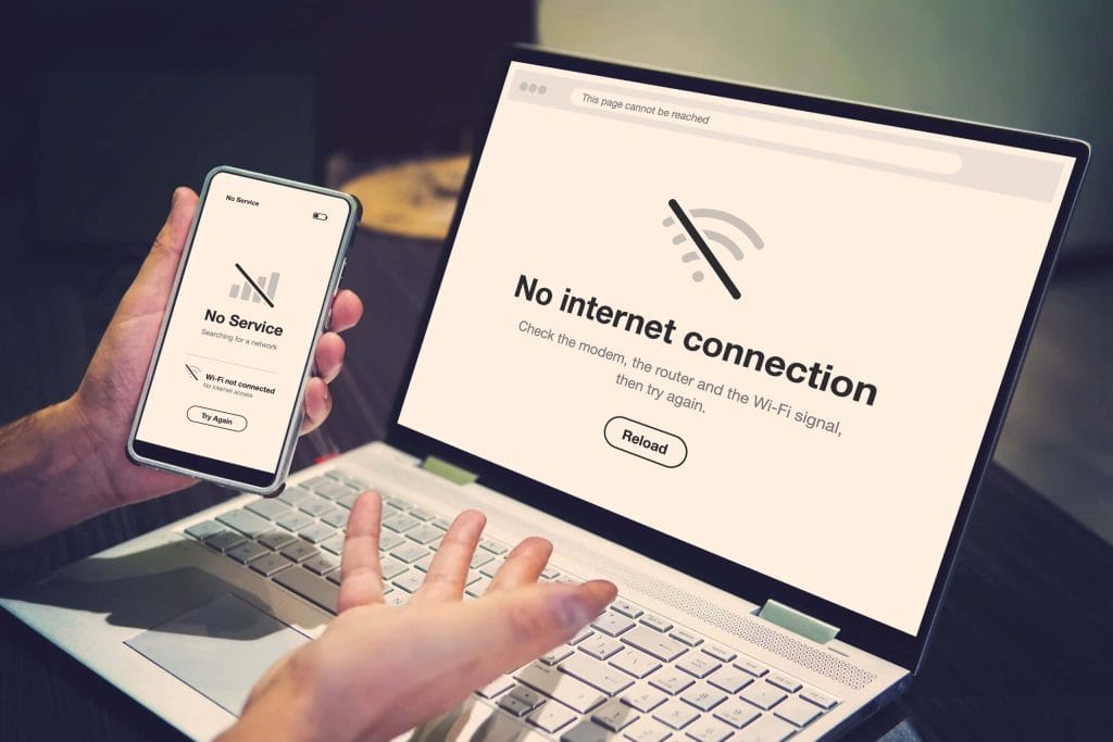 A person's hands holding a smartphone that shows a No Service screen, in front of an open laptop showing a No internet connection page.