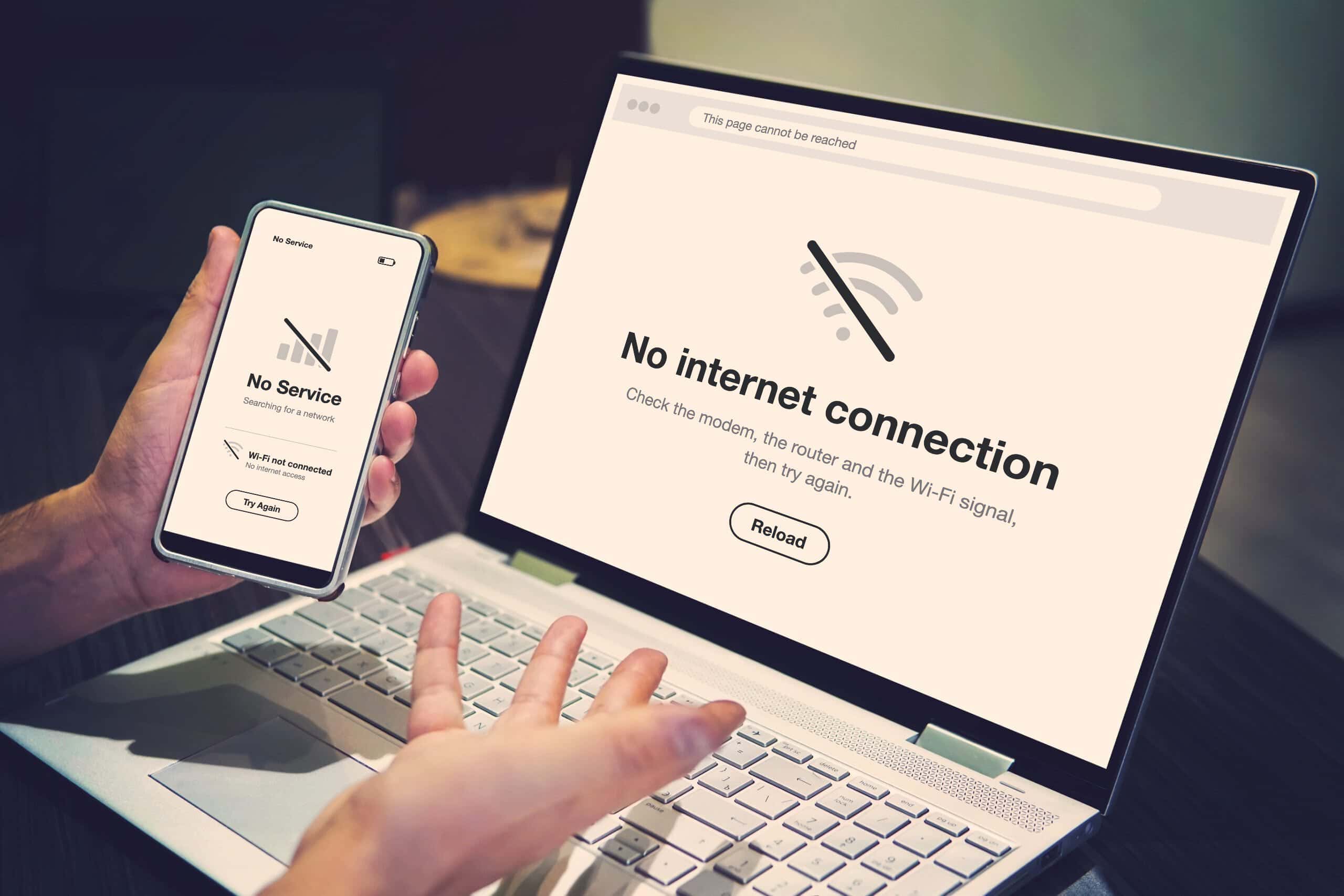 A person's hands holding a smartphone that shows a No Service screen, in front of an open laptop showing a No internet connection page.
