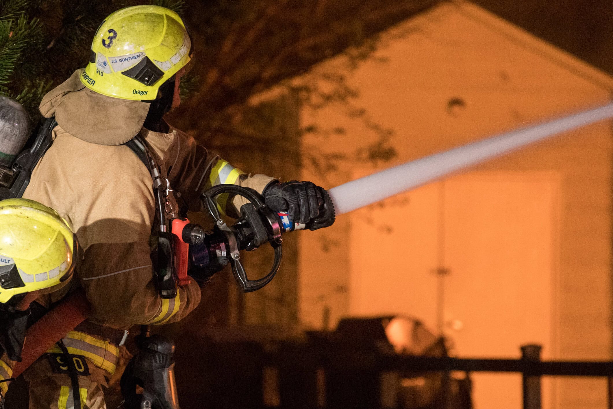 A firefighter directing a hose toward a house as brush burns nearby in California