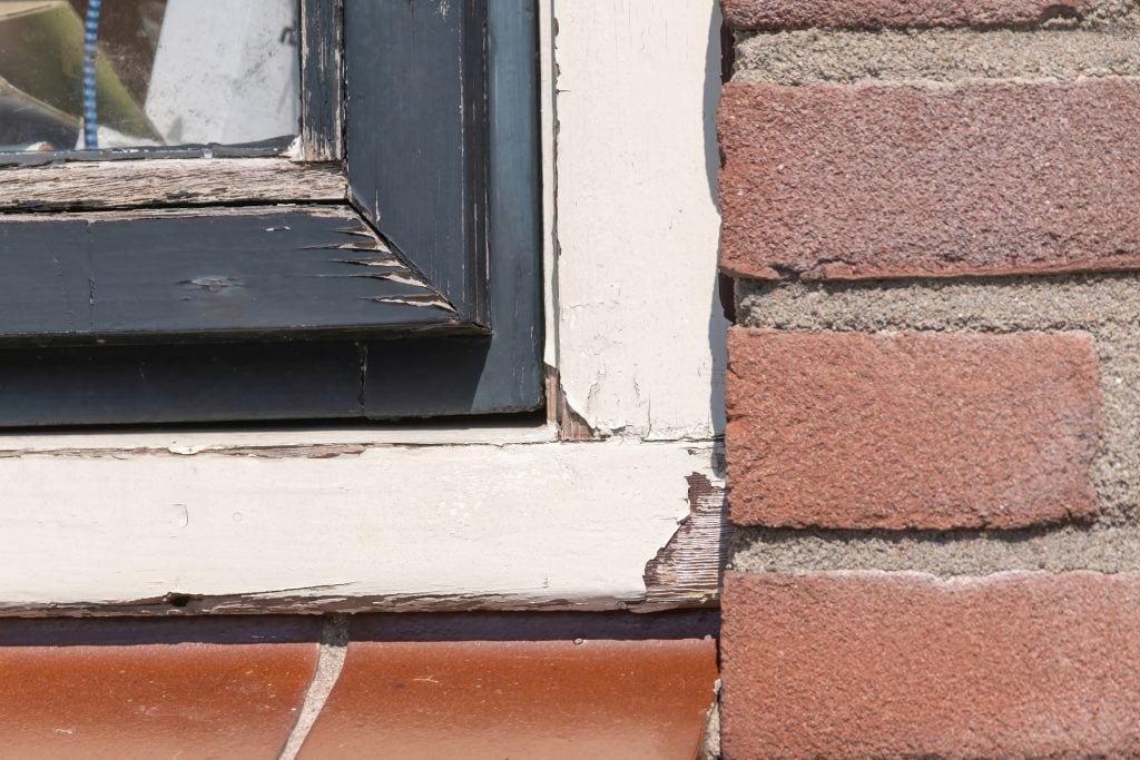 Close-up of a window frame with peeling black and cream paint, a worn wooden sill, and a red brick wall beside it.