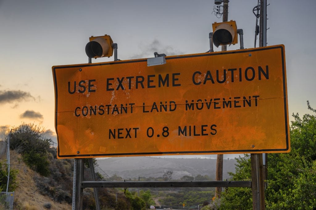 An orange road sign reading Use Extreme Caution, Constant Land Movement, Next 0.8 Miles, with two warning lights above it and hillside brush behind