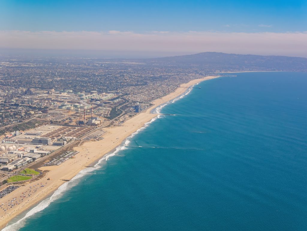 Aerial view over El Segundo Beach, with surf breaking along a wide sand shoreline, the city's low-rise street grid inland, and a headland in the far distance.