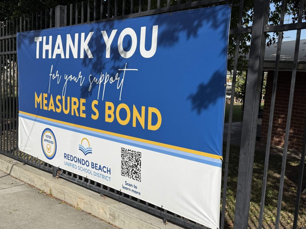 Blue and white vinyl banner reading Thank You for your support, Measure S Bond, with a Voter Approved seal, the Redondo Beach Unified School District logo and a QR code, hung on a black iron fence.