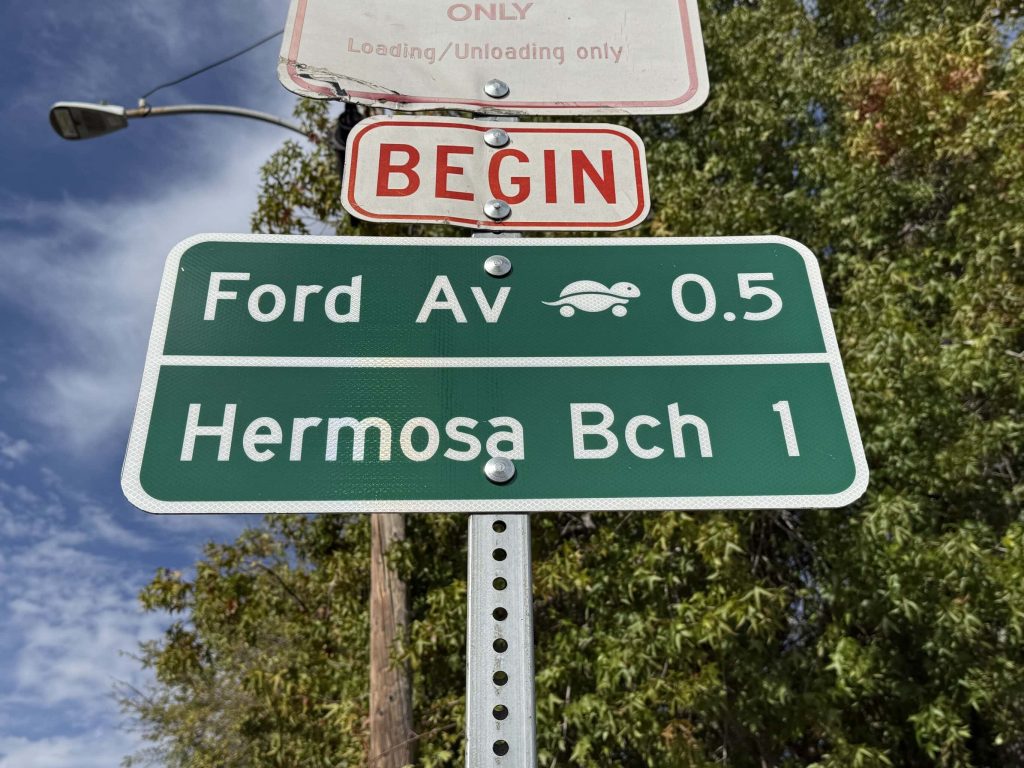 Green street sign reading Ford Av 0.5 with a rolling turtle symbol above Hermosa Bch 1, mounted under a red BEGIN plate and a loading and unloading only sign.